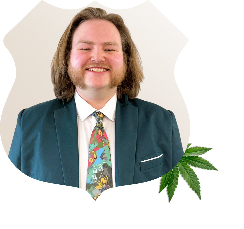 Connor Phillips – Des Peres Store Manager Portrait of Connor Phillips, store manager at Root 66 Dispensary Des Peres. He has long brown hair. A cannabis leaf is in the bottom corner of the image.